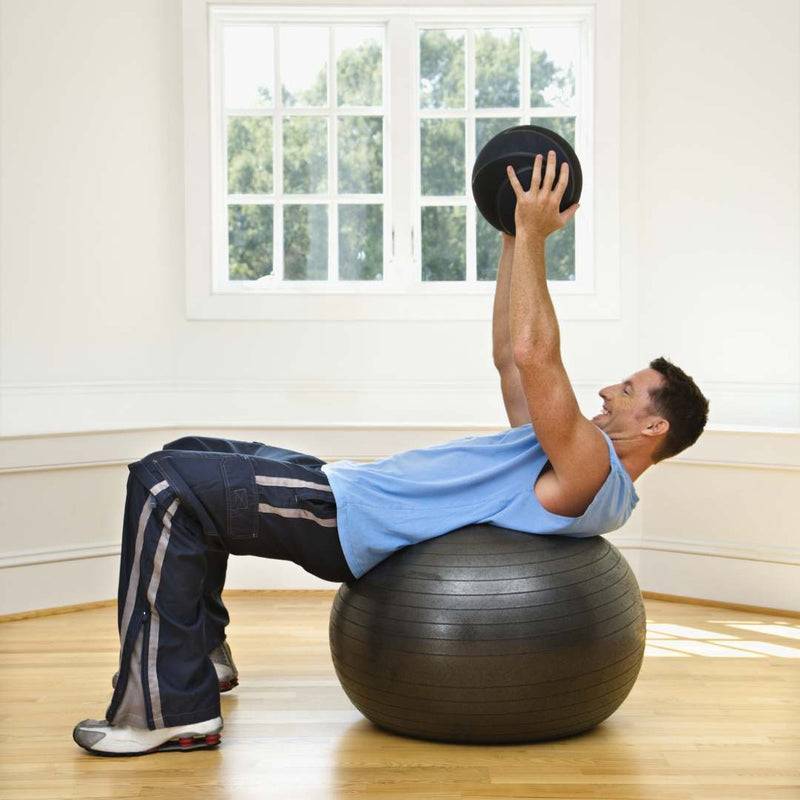 A man in a light blue tank top and dark track pants performs a crunch exercise on a black stability ball, holding a black medicine ball overhead in a bright room with wood floors and a white window.