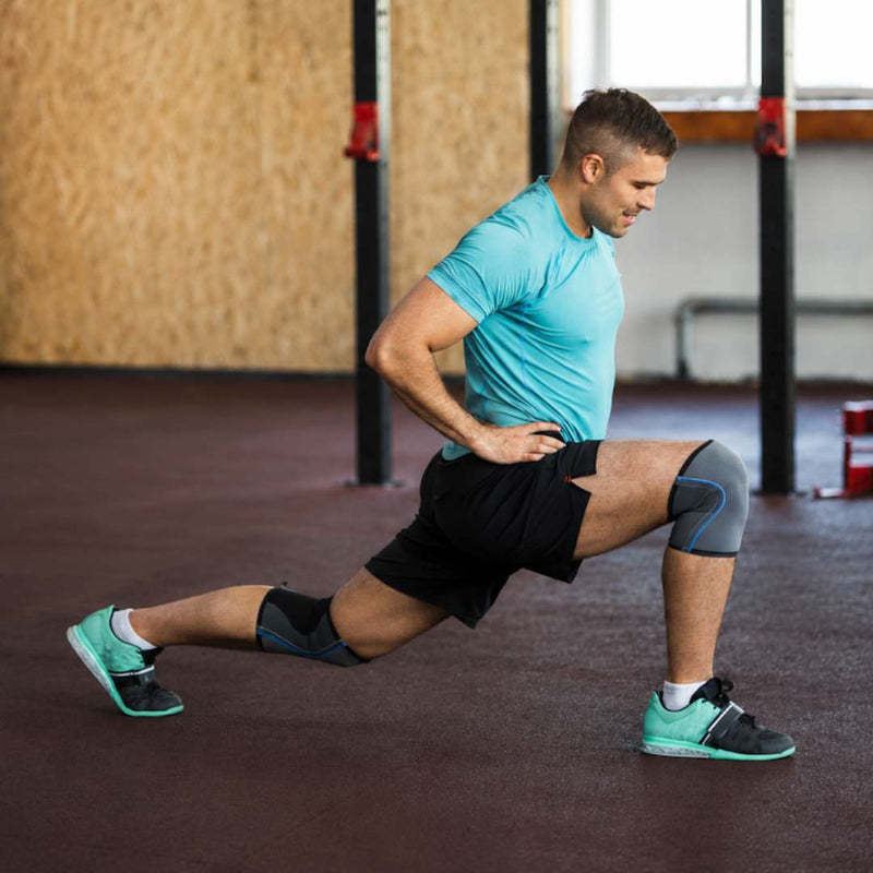 A man in a gym performing a lunge stretch, wearing a light blue t-shirt, black athletic shorts, and teal and black running shoes with knee support sleeves.