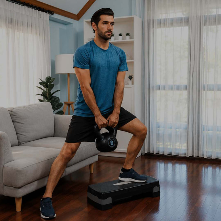 A man in a blue t-shirt and black shorts performs a kettlebell squat while stepping onto a black platform in a modern living room with a gray sofa, white curtains, and wooden floors.