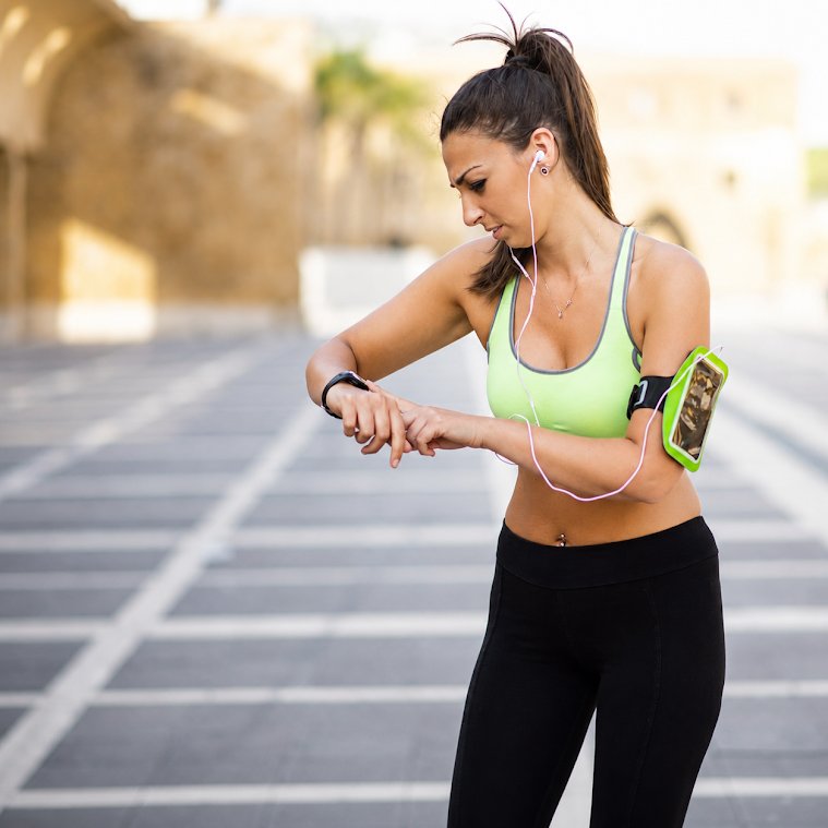 A woman in athletic wear, featuring a neon green sports bra and black leggings, checks her smartwatch outdoors on a paved path. She has a green and black armband phone case on her arm and is wearing white earphones.